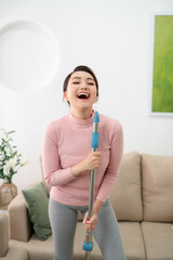 Happy young woman singing while washing floor at living room, using mop as microphone