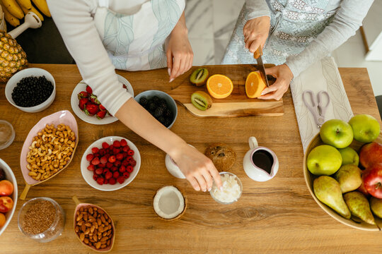 View From Above Of Women Hands Cooking At Home, Cutting Orange Fruit With Knife With Nuts And Seeds, Berries And Fruits On Wooden Working Surface At Kitchen.