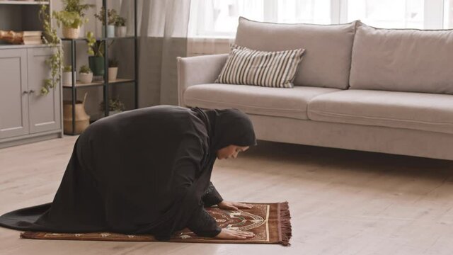 Side-view shot of young Muslim woman in traditional black clothing and hijab praying on floor in living room