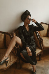 Obraz premium Fashionable lady in long sleeve dress and sunglasses posing inside. Modern woman in heels and hat sitting on armchair in apartment..