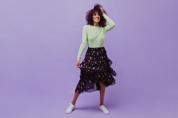 Pretty cool brunette woman in midi black skirt and green top smiles on purple background. Happy curly girl moves on isolated.