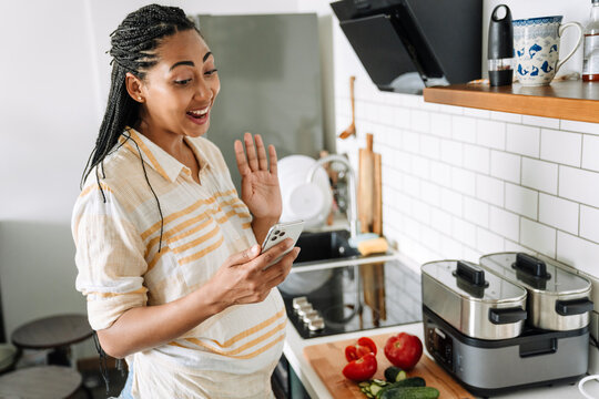 Black pregnant woman gesturing and using mobile phone in kitchen