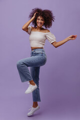 Cheerful curly brunette woman in denim pants and white top dances on purple background. Attractive dark-skinned girl smiles.