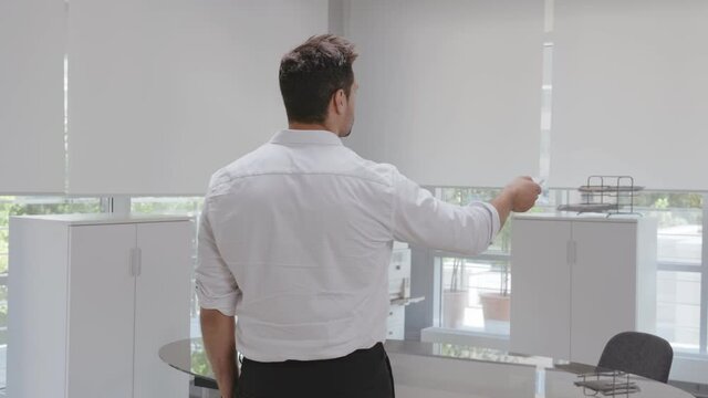 Rear view of young businessman open window blinds with remote control