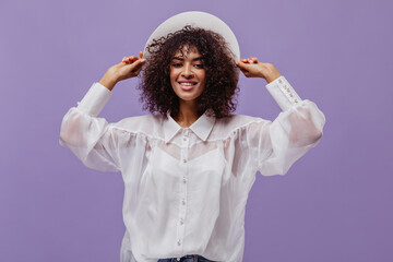 Portrait of brunette woman in white blouse and hat. Attractive dark-skinned curly lady smiles on...