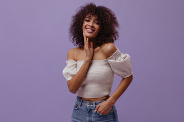 Dark-skinned woman smiles on purple background. Cheerful curly lady in white tip looks into camera...