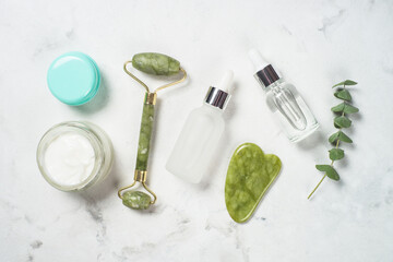 Jade roller and gua sha stone massager with face cream and serum bottle. Trendy cosmetic products at white. Anti aging therapy. Top view.
