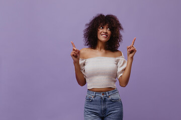 Charming brunette woman points up at place for text on purple background. Happy dark-skinned curly...