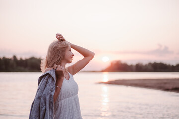 Obraz premium Portrait in profile of a young woman on the beach. A beautiful blonde in a white summer dress stands on the river bank and holds a denim jacket in her hand against the background of sunset and water.