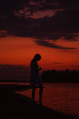 Fototapeta premium The silhouette of a woman in profile against the background of the sunset. A slender young woman walks along the river bank against the background of clouds and the beach. 