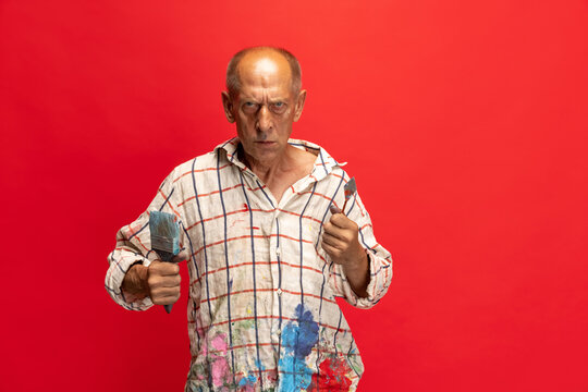 Mature or senior grey painter with a brush standing in front of camera. Funny man in a paint-stained shirt fooling around isolated on red