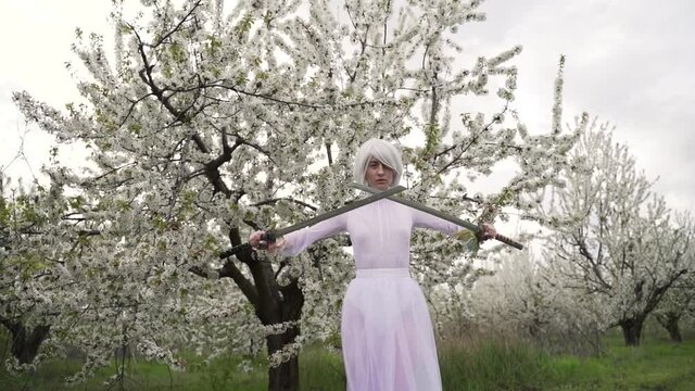 Blonde bride in white dress holds sharp metal swords in her hands. Young woman in white clothes exercises with fencing in the flowering gardens in the spring. Cosplay 