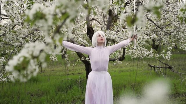 Young woman with wig in white dress trains with sharp sword in the blooming gardens in spring