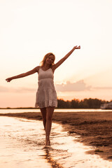 Beautiful woman posing on the beach. A slender young blonde in a white dress is spinning on the riverbank against the sunset background and raises her hands up. 