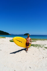 A man stands on a sandy shore and holds a large yellow paddle board.