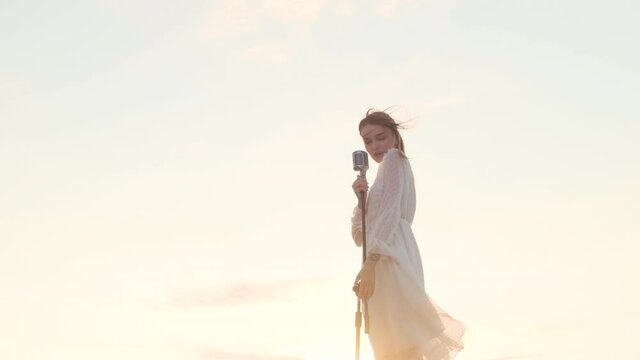beautiful woman in white dress on top of mountain, on edge of abyss and sings song into shiny retro vintage microphone. Attractive lady singer on background of sunset or dawn sky. Sun shine in camera.