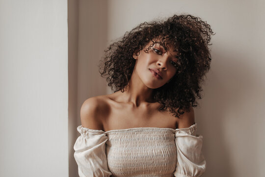 Cheerful curly woman in white cropped top looks into camera. Dark-skinned brunette lady poses in room with white walls.