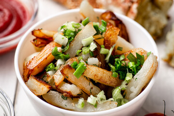 Fried potatoes in a white plate with green onions, parsley, tomatoes on a white wooden background.