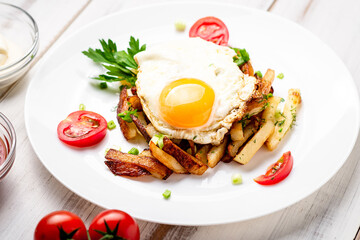 Fried potatoes with egg, herbs and tomatoes on a white plate on a white wooden background.