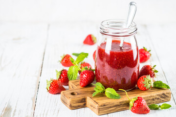 Strawberry jam in the glass jar at white table with fresh strawberries.