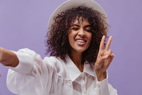 Attractive curly brunette woman in white blouse and hat takes selfie, winks and shows peace sign on purple background.