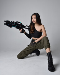 Full length portrait of pretty brunette, asian girl wearing black top and khaki utilitarian army pants and leather boots. Sitting pose holding a science fiction gun, isolated agent a light grey studio