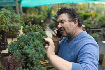 Caucasian male gardener talking by smartphone and touching bonsai tree at garden centre