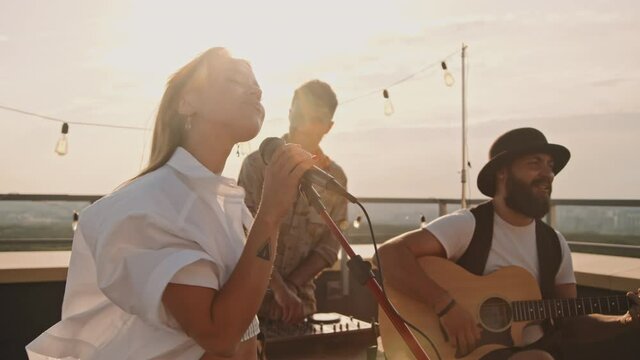 Slowmo tracking shot of band performing song on rooftop terrace on summer evening. Young woman singing into microphone as men DJing and playing acoustic guitar