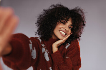 Charming dark-skinned lady takes selfie on isolated. Happy curly brunette woman in burgundy sweater smiles on grey background.