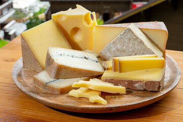 Cheese collection, wooden board with French cheeses comte, beaufort, abondance, emmental, morbier and french mountains village in Haute-Savoie on background