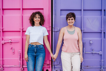 happy lesbian couple smiling, looking at camera and holding hands over pink and purple background. Love is love. LGTBI concept - obrazy, fototapety, plakaty