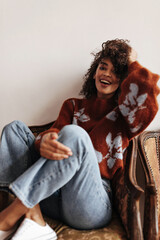Excited dark-skinned woman in jeans, burgundy sweater ruffles hair and smiles sincerely sitting on...