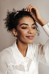 Charming brunette curly woman in white blouse looks away. Portrait of attractive dark-skinned lady smiling on grey background.