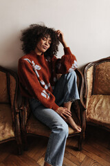 Calm brunette woman sits on chair. Dark-skinned lady in jeans and warm burgundy sweater poses with...