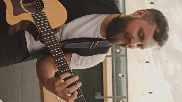 Handheld vertical shot of happy bearded young man playing acoustic guitar on rooftop terrace on nice summer evening