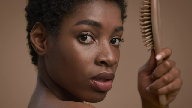 African American Woman Brushing Short Hair Holding Hairbrush, Beige Background