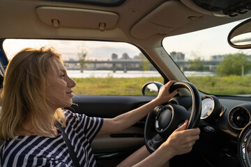 Naklejka premium Happy smiling woman driving modern vehicle. Caucasian female sitting alone on driver seat with fasten belt. Attractive confident blonde travelling by car. Lifestyle and success concept