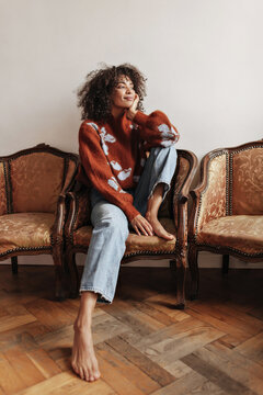 Beautiful dark-skinned woman in denim pants and warm sweater sits on chair. Curly brunette girl relaxes and leans on knees.