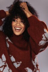 Dark-skinned curly woman ruffles hair on grey background. Cheerful girl in warm red sweater smiles...