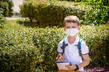One pupil boy wearing mask is sitting on bench near his school during covid-19 pandemic time. New...