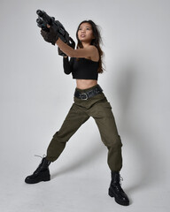 Full length portrait of pretty brunette, asian girl wearing black top and khaki utilitarian army pants and leather boots. Standing pose holding a science fiction gun, isolated agent a light grey studi