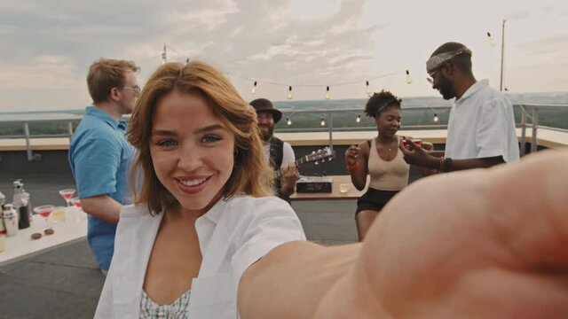 POV shot of happy young woman holding camera and taking selfie or filming during rooftop party with her friends. Cheerful man playing acoustic guitar
