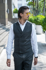 Portrait of handsome young man in black formal suit and yellow sunglasses walking on the street and looking aside