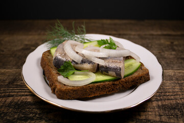 A sandwich of rye bread with salted herring on a white plate.A delicious snack on a white plate with fresh herbs on a wooden table.