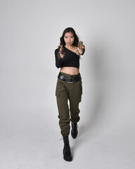 Full length portrait of pretty brunette, asian girl wearing black top and khaki utilitarian army pants and leather boots. Standing pose holding a science fiction gun, isolated agent a light grey studi