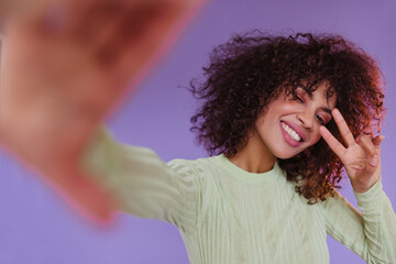 Happy curly woman takes selfie on purple background. Charming dark-skinned brunette lady shows peace sign and smiles.