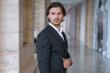 Stylish young brunet man in black suit standing on the street near modern office building. Portrait.