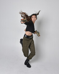Full length portrait of pretty brunette, asian girl wearing black top and khaki utilitarian army pants and leather boots. Standing pose holding a science fiction gun, isolated agent a light grey studi