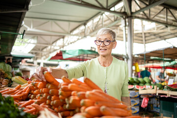 Portrait of senior caucasian woman buying fresh organic vegetables and fruit at market place and holding bag full of healthy food.