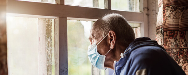 Older man wearing face mask looking through window with sorrow